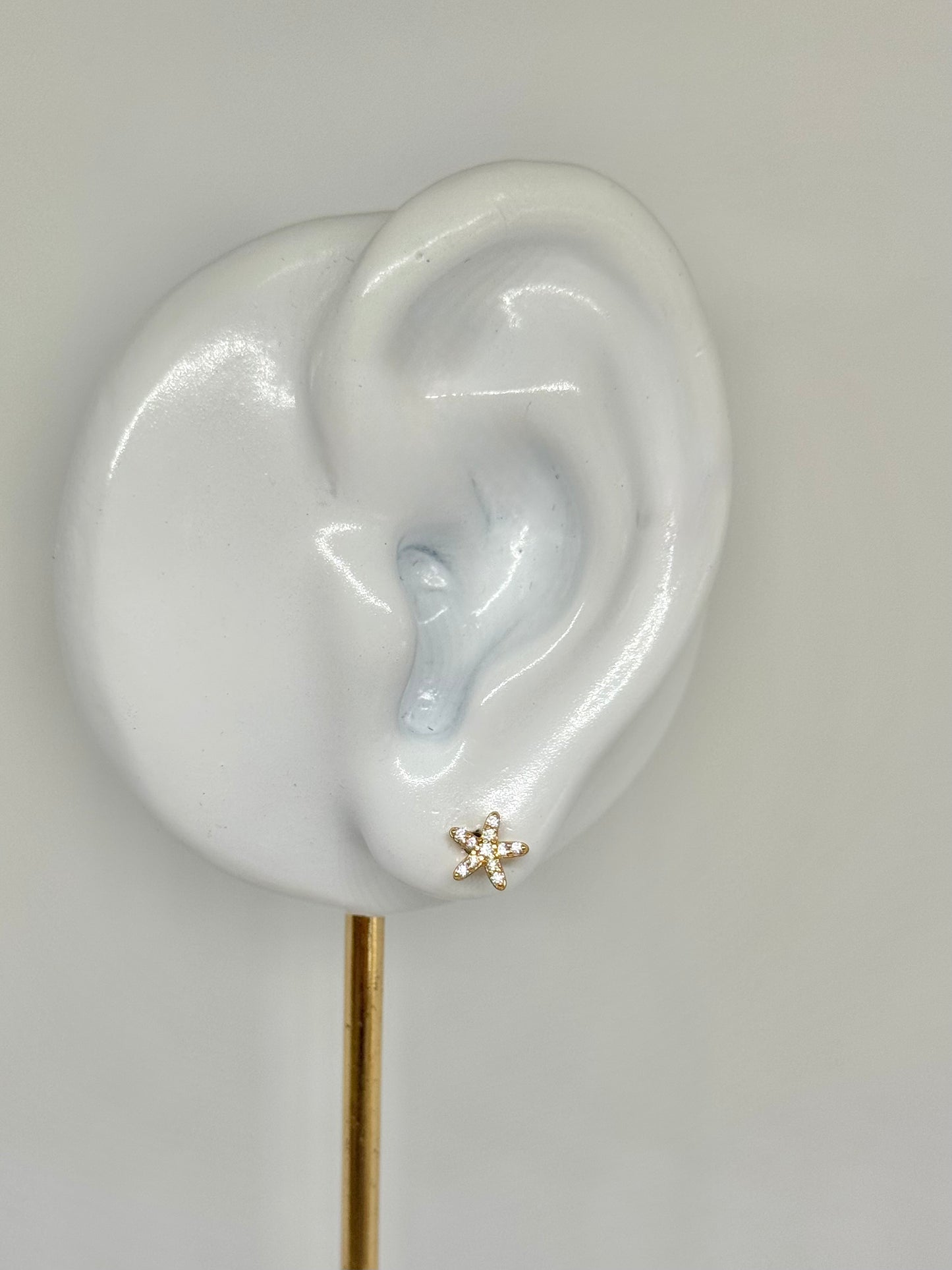 Close-up of an ear wearing a gold earring with a clear gemstone on a plain background, star earrings, gemstone, everyday wear, stud earrings