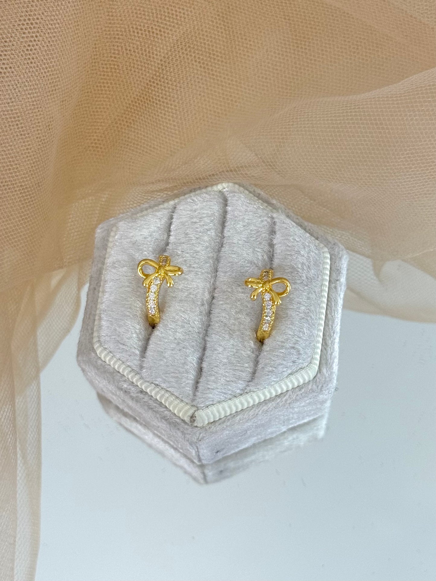 Gold bow-shaped earrings in a hexagonal gray velvet box on a beige fabric background, bow, bow earrings, bow stud earrings, perfect gift for her, modern design earrings, minimalist look, classic look, elegance, everyday wear