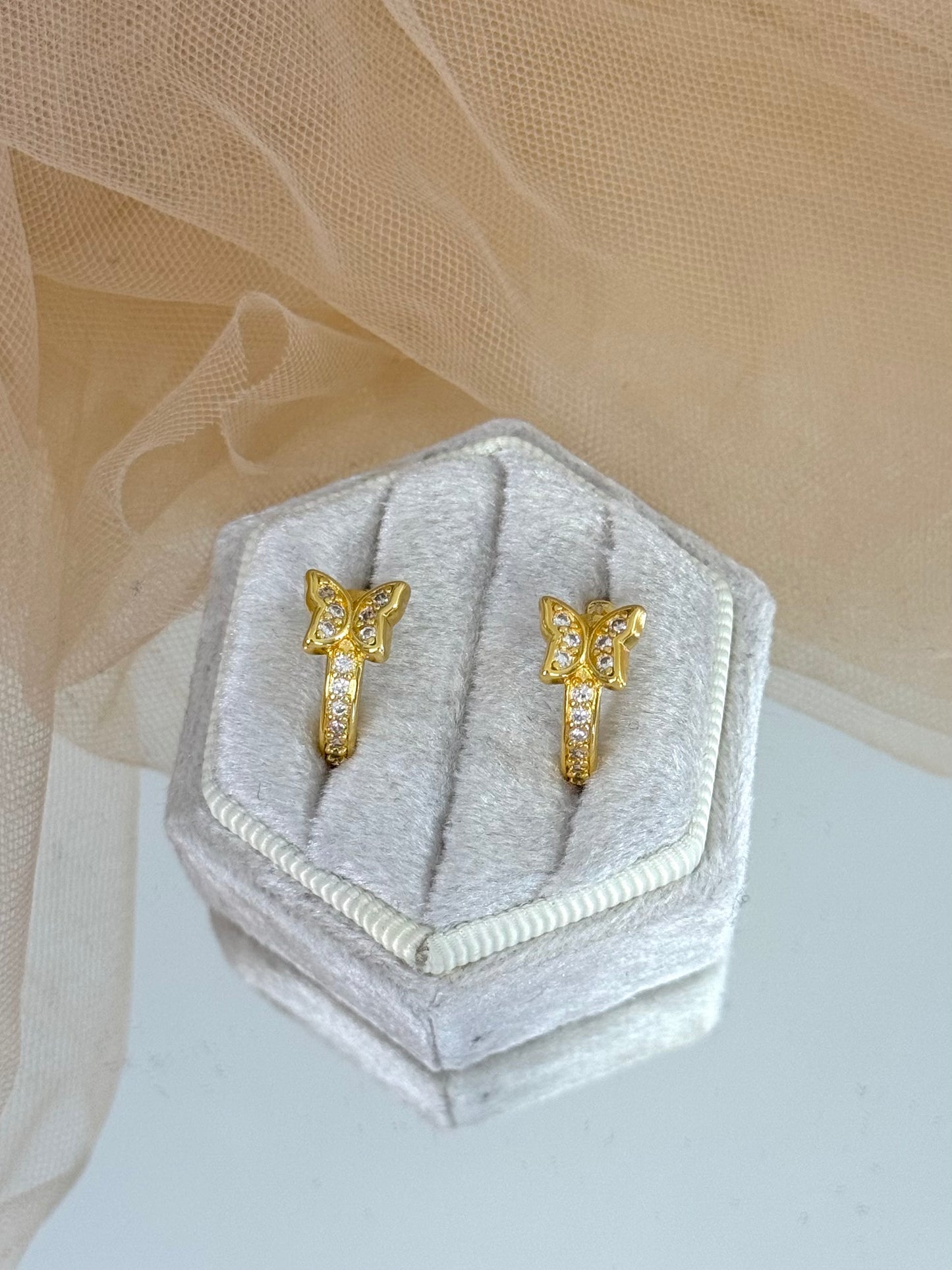 Gold butterfly-shaped earring on a hexagonal gray velvet stand with a beige fabric background, butterfly, butterfly stud earrings, elegance, everyday wear