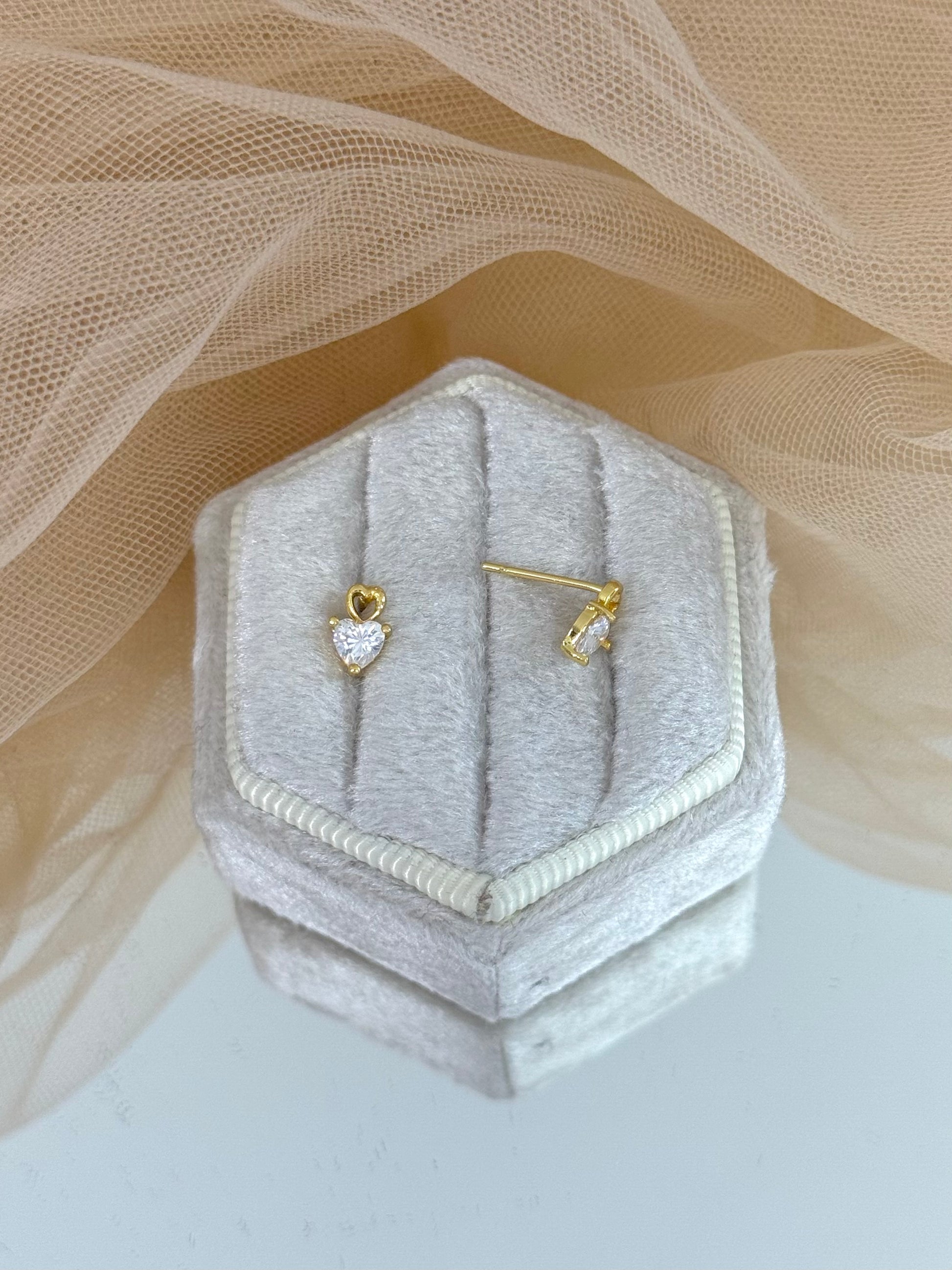 Gold earrings with heart-shaped gemstones in a hexagonal gray velvet box on a beige fabric background, heart shaped, heart stud earrings, stud earrings, minimalist yellow gold, everyday wear