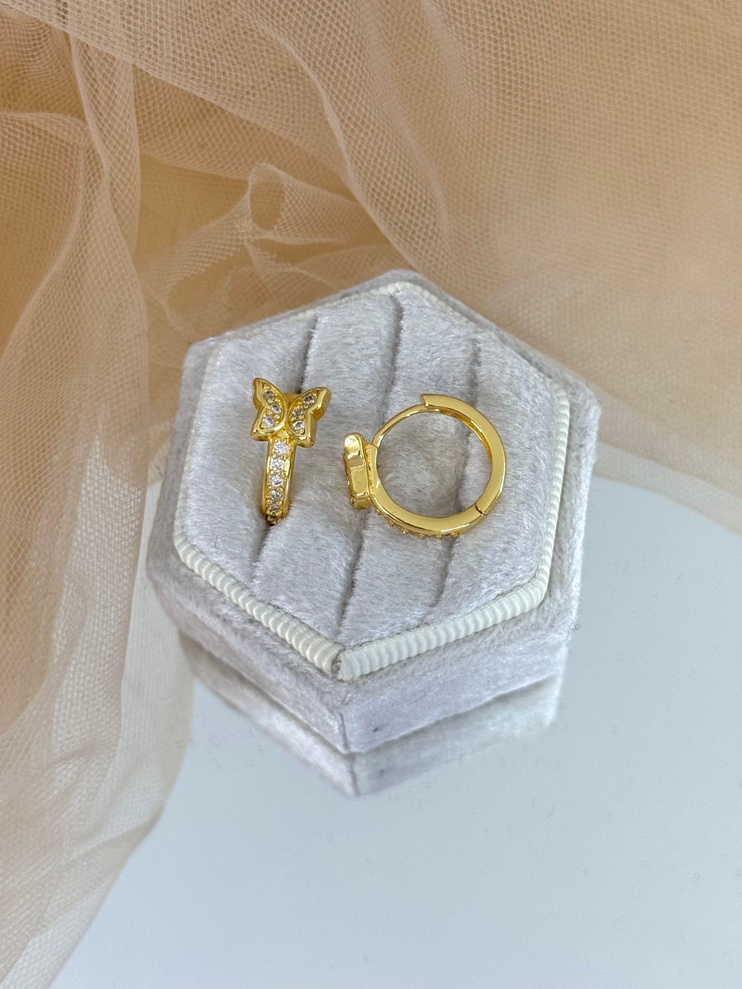 Gold butterfly-shaped earring on a hexagonal gray velvet stand with a beige fabric background, butterfly, butterfly stud earrings, elegance, everyday wear
