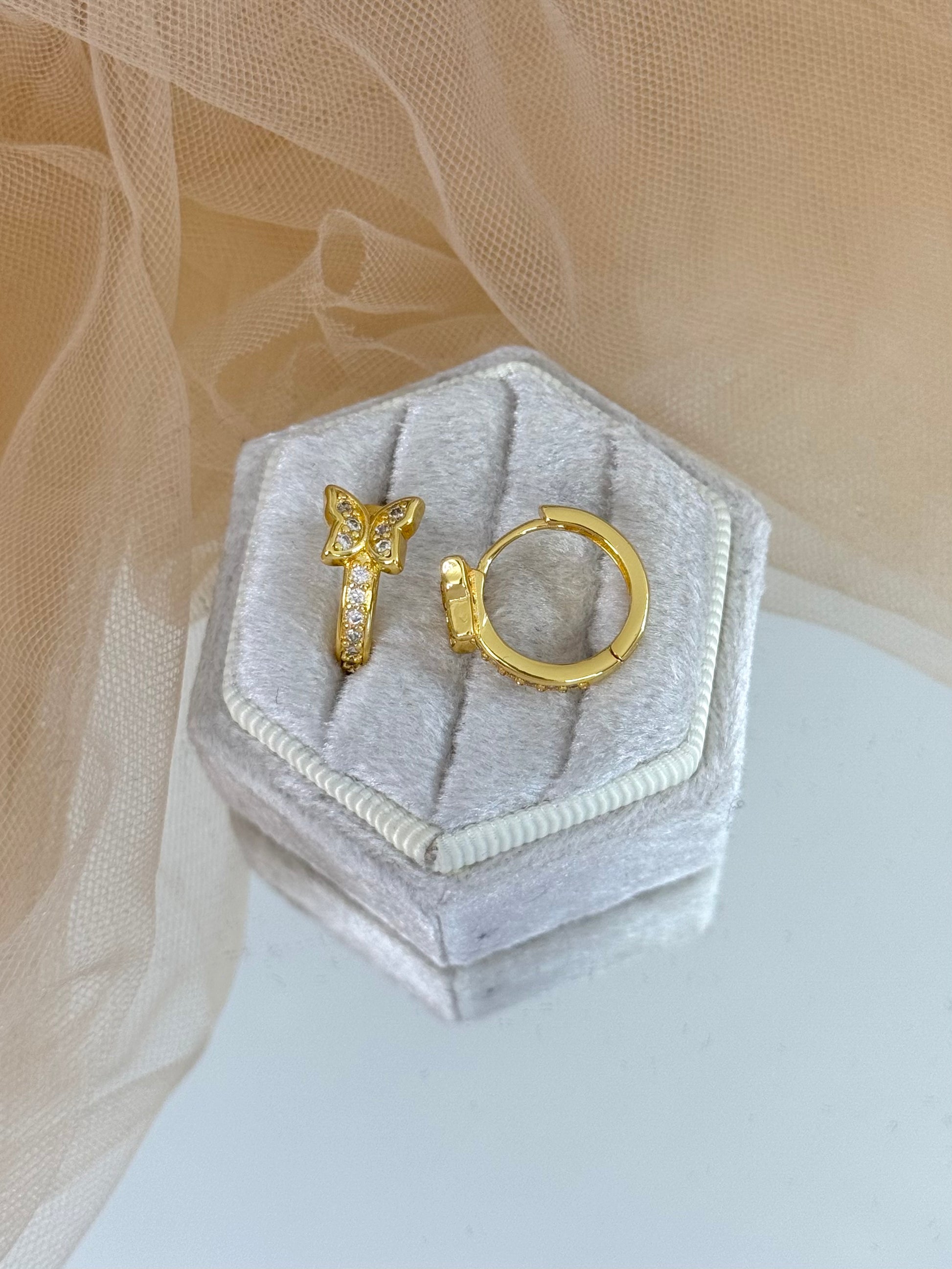Gold butterfly-shaped earring on a hexagonal gray velvet stand with a beige fabric background, butterfly, butterfly stud earrings, elegance, everyday wear